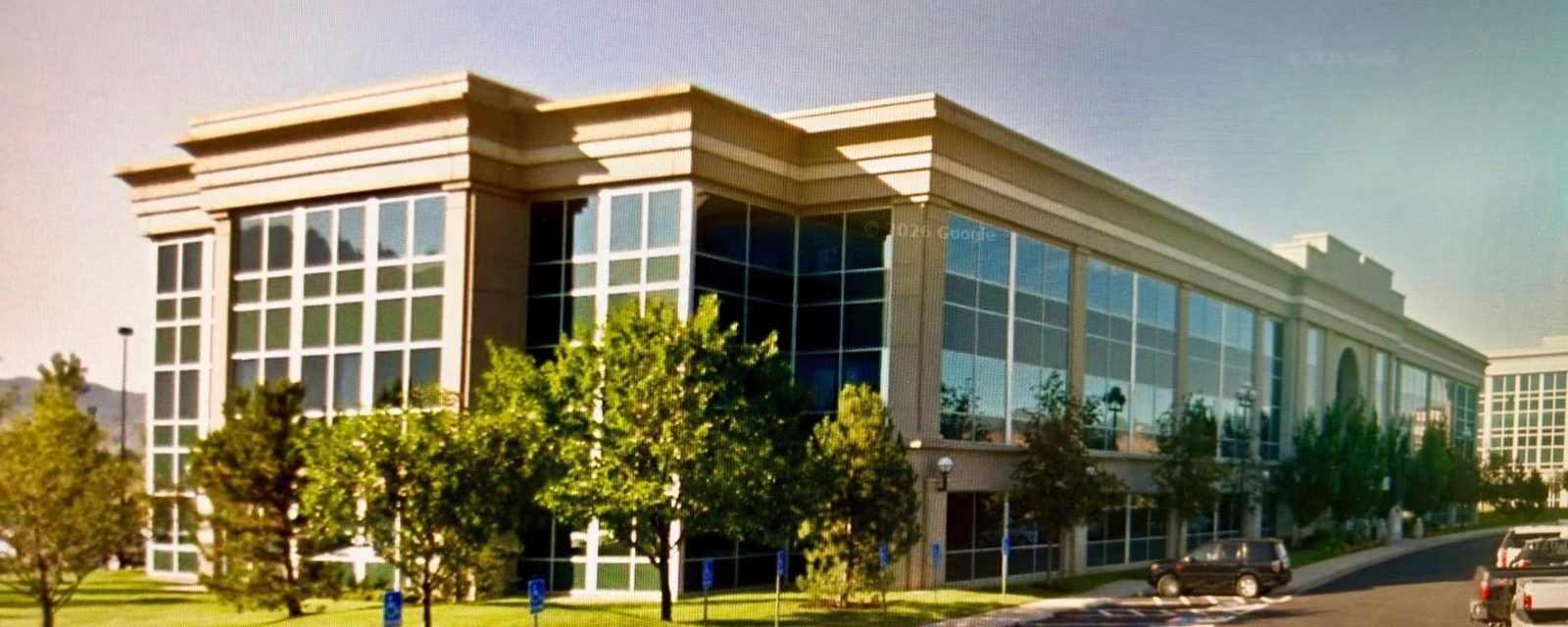 A multi-story glass and stone office building with trees and visitor parking in front.